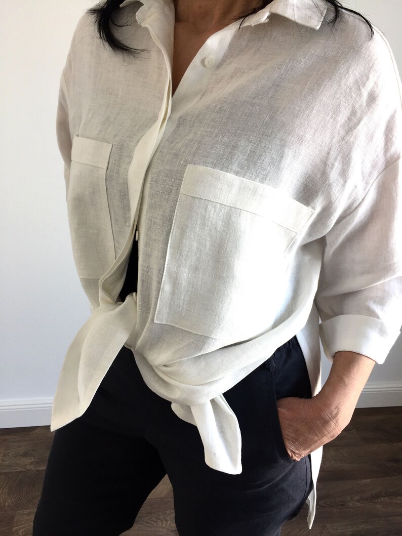 May include: A white linen shirt with long sleeves and two large pockets. The shirt is tied at the waist and worn with black pants.
