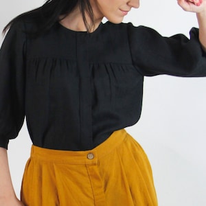 Puede incluir: Una blusa negra de manga larga con cuello fruncido y manga ligeramente abullonada. La blusa se combina con una falda amarilla mostaza.