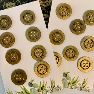 May include: A collection of fourteen circular buttons with a metallic gold finish, each featuring four holes in the centre. The buttons are arranged on two white rectangular display cards, each decorated with a green botanical leaf illustration.
