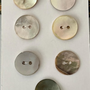 May include: Seven circular mother-of-pearl buttons arranged on a white card with the text 15 MM printed at the top. Each button features two central holes and displays unique iridescent colour variations ranging from cream to grey and brown.