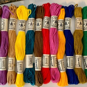 May include: A selection of embroidery threads in a variety of colours, such as red, pink, yellow, and blue. Each skein is labelled with a number and the brand name. The threads are neatly arranged, displaying a spectrum of shades for craft projects.