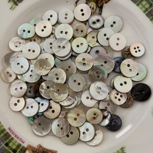 May include: A collection of round, iridescent mother-of-pearl buttons in various shades of white, cream, and grey. Each button features two or four holes in the centre. The buttons are piled together on a white plate with a tartan pattern.