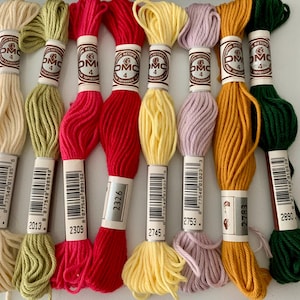 May include: A selection of DMC embroidery threads in a range of colours: cream, green, pink, red, yellow, lavender, gold, and dark green. Each skein is labelled with a number and the DMC logo. The threads are tightly wound and ready to use.