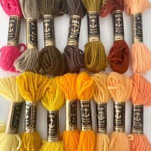 May include: A selection of Anchor Tapisserie Wool-Laine yarn skeins in shades of pink, beige, brown, yellow, orange, and rust. Each skein is labelled with a number and the Anchor brand logo.