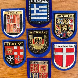 Peut inclure: Collection d'écussons brodés en forme de boucliers, représentant différents pays. Les écussons ont des bordures bleues et présentent des drapeaux et symboles nationaux. Les pays incluent la Grèce, l'Espagne, l'Italie, l'Allemagne, la Belgique, le Danemark et la Grande-Bretagne.
