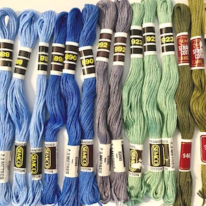 May include: A collection of thirteen skeins of Semco stranded cotton embroidery thread arranged in a gradient from light blue to deep blue, purple, light green, and olive green. Each skein features a paper label with the colour code and 7.3 metres.