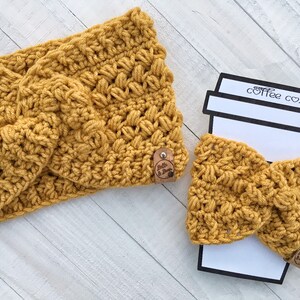 May include: A set of two crocheted items in a mustard yellow color. The larger item is a headband and the smaller item is a coffee cozy. Both items have a knotted design and feature a small brown leather tag with the text "Belle & Bloom".