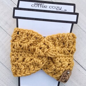 May include: A yellow crocheted coffee cozy with a brown leather tag that says "Belle & Bean". The cozy is displayed on a white coffee cup cutout with the words "coffee cozy" written on it.