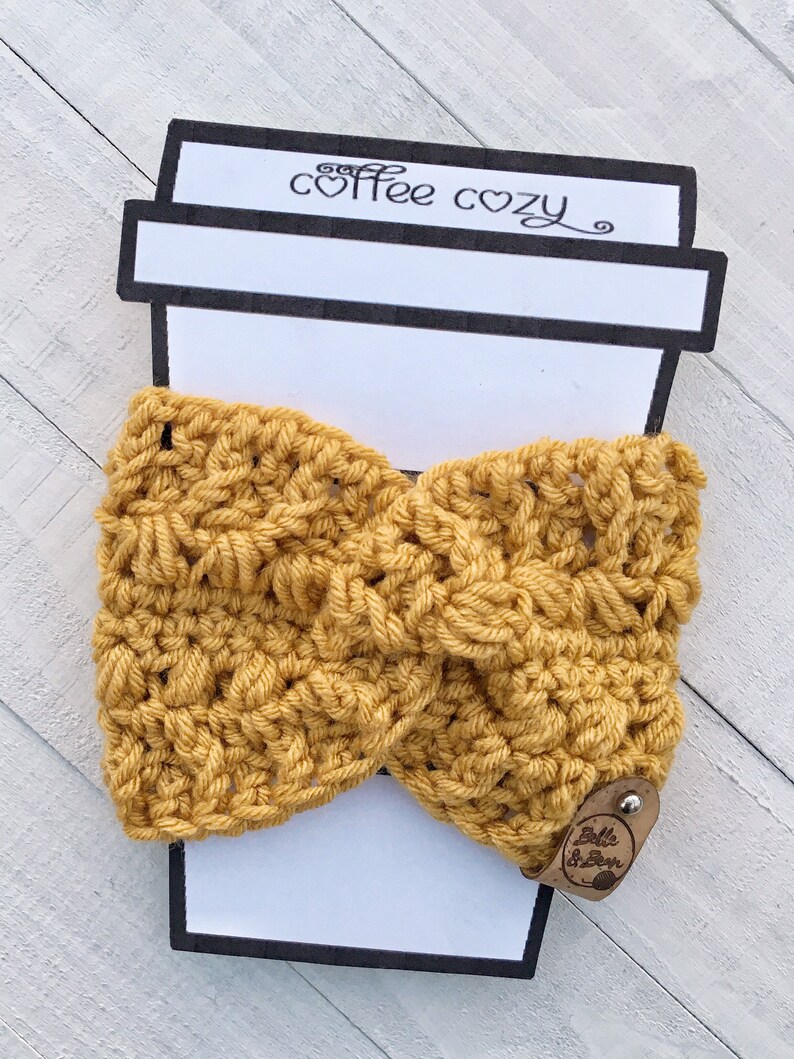 May include: A yellow crocheted coffee cozy with a brown leather tag that says "Belle & Bean". The cozy is displayed on a white coffee cup cutout with the words "coffee cozy" written on it.