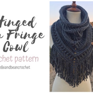 May include: A gray crochet cowl with fringe on a mannequin. The cowl has a textured pattern and a small brown button on the side. The text "Hinged On Fringe Cowl crochet pattern @belleandbeancrochet" is visible in the image.