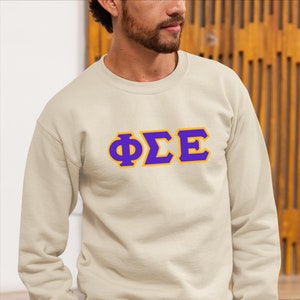 May include: A beige sweatshirt with a purple and yellow applique of the Greek letters "ΦΣΕ". The applique has a zig-zag stitch around the edges.