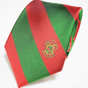 May include: A red and green striped tie with a gold embroidered crest. The crest features a shield with a lion and a crown.
