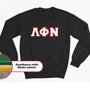 Könnte beinhalten: Schwarzes Sweatshirt mit weißen und roten griechischen Buchstaben ΛΦΝ. Das Sweatshirt zeigt den Text "Applikation mit Satinstich" in einem weißen Oval. Eine Nahaufnahme zeigt die Stichdetails in Grün, Weiß, Gelb und Grau.