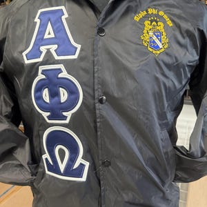 May include: Black nylon jacket with the Greek letters Alpha, Phi, and Omega in blue with white trim. The jacket also features a crest with the words "Alpha Phi Omega" in gold.