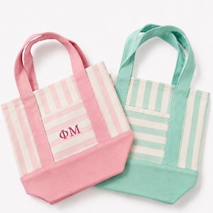 May include: Two small canvas tote bags with vertical stripes. One bag is pink and white with the Greek letters ΦΜ embroidered in pink on the front pocket. The second bag is mint green and white with a matching mint green base and two top handles.
