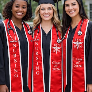 May include: Three smiling graduates wearing black caps and gowns with red and white nursing stoles. The stoles have the word "NURSING" vertically and "BSN RN Class of 2026" printed on them. Each graduate has a stethoscope around their neck.
