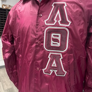 Puede incluir: Chaqueta de nailon color burdeos con cierre de botones y cuello clásico. La chaqueta presenta las letras "AOA" en un diseño de estilo universitario blanco y burdeos en la parte delantera. Una prenda de abrigo informal.