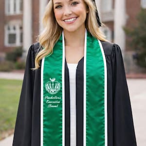 Può includere: Una persona indossa una toga di laurea nera e un tocco. Una fascia verde e bianca con la scritta "Panhellenic Executive Board" è drappeggiata sulla toga. Lo sfondo presenta un edificio in mattoni.