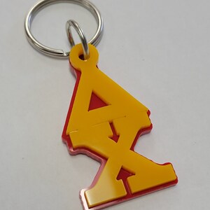 Puede incluir: Un llavero rojo y amarillo con un diseño estilizado de la letra 'X'. El llavero tiene un anillo de metal plateado.