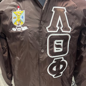 May include: A brown nylon jacket with a button-up front. The jacket has a crest on the left chest and the Greek letters ΛΟΦ in a varsity style on the right chest. The letters are brown with white trim.
