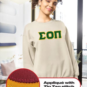 May include: A beige sweatshirt with a green and yellow Greek letter design that says "ΣΟΠ" on the front. The sweatshirt has a zig-zag stitch around the design.