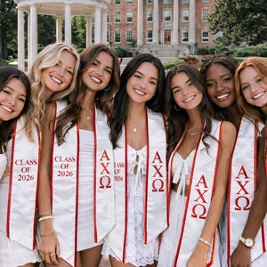 May include: Group of seven women smiling, wearing white dresses and graduation sashes. The sashes are white with red trim and feature Greek letters and "Class of 2026". The background includes a university building and a gazebo.