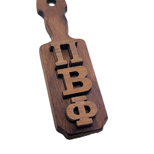 May include: A wooden paddle with the Greek letters ΠΒΦ carved into it. The paddle is a dark brown color and has a hole at the top for hanging. The letters are raised and in a lighter shade of brown.