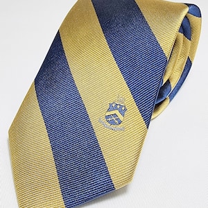 May include: A gold and navy blue striped tie with a crest featuring a shield with a crown and stars. The crest is embroidered on the tie.