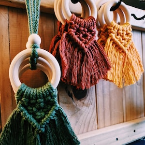 Puede incluir: Tres mordedores de anillo de madera con flecos de macramé de colores. Los mordedores son verde, rojo y amarillo.