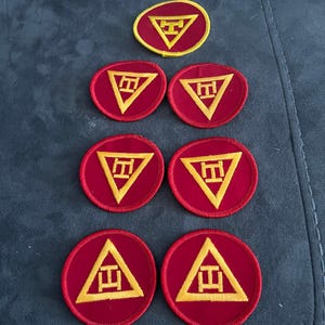 May include: A collection of embroidered patches. The patches are primarily red with a yellow geometric design. The design features a yellow triangle with a stylized letter or symbol inside. The patches are in various shapes, including a circle and an oval.