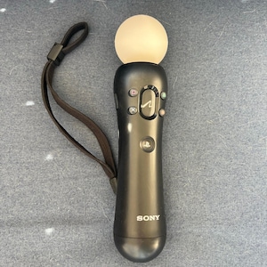 May include: A black Sony PlayStation Move motion controller with a white orb at the top. The controller has several buttons and a wrist strap. The word "SONY" is printed on the controller.
