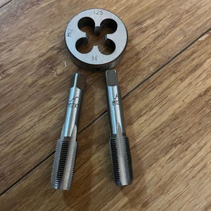 May include: A set of metal tools for threading, including a die and two taps. The die is round with four holes and markings including "1.25" and "M12". The taps are cylindrical with threaded sections and "M12X1" and "M12" markings.