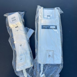 May include: Two white Aiphone intercom units, one a handset and the other a base station, both sealed in clear plastic. The handset has a speaker and buttons, while the base station features the Aiphone logo and a rectangular opening.