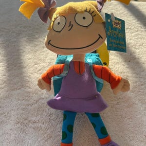 May include: A plush Angelica doll from the Rugrats animated series. The doll features yellow hair, a purple dress, blue leggings with green polka dots, and orange sleeves. It has a backpack and a tag that reads "Angelica's Backpack Book".
