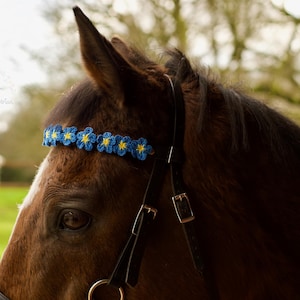 May include: A brown horse with a blue and yellow floral headband and a black bridle. The image is a close-up of the horse's head, with a blurred green and brown background. The headband is made of small, crocheted flowers.