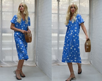 Puede incluir: Una mujer lleva un vestido midi azul con estampado floral blanco y un bolso de paja. Lleva sandalias de tacón negras.