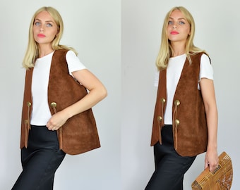 May include: A brown suede vest with fringe and gold button accents. The vest is worn over a white t-shirt and black skirt.