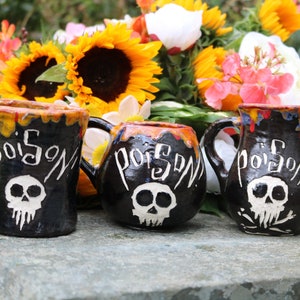 Könnte beinhalten: Drei kleine schwarze Keramikbecher mit weißen Totenkopf und Knochen und dem Wort "Gift" darauf. Die Becher stehen auf einer grauen Steinoberfläche vor einem Strauß Sonnenblumen und weißen Blumen.
