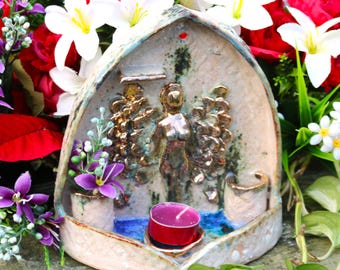 May include: A ceramic wall hanging with a sculpted angel figure with gold wings. The angel is standing in a small alcove with a teal blue pool of water and a red candle holder. The wall hanging is decorated with a white and brown glaze.