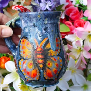Op de afbeelding: Een blauwe keramische mok met een geschilderde vlinder aan de voorkant. De vlinder heeft oranje, gele en rode vleugels. De mok heeft een handvat in de vorm van een paddenstoel.