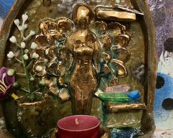 May include: A ceramic wall hanging with a gold-colored female figure, a small plant, and a tealight candle holder. The figure is surrounded by a green and blue floral design. The wall hanging has a hole at the top for hanging.