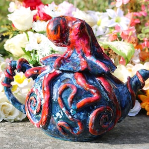 May include: A ceramic teapot with a blue base and red octopus design. The teapot features a lid with an octopus figure and red tentacles wrapping around the blue body. The handle and spout are also adorned with red accents.
