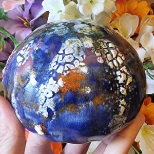 Pode incluir: Uma esfera de cerâmica azul com esmalte craquelado branco e laranja. A esfera tem uma superfície texturizada e é pintada à mão.