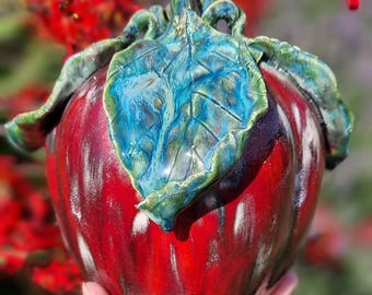 Può includere: Una scultura di mela in ceramica con una finitura rosso intenso lucida e una foglia verde e blu strutturata e prominente attaccata alla parte superiore. La superficie della mela presenta creste verticali e sottili riflessi bianchi, mentre la foglia mostra venature complesse e una smaltatura multicolore.
