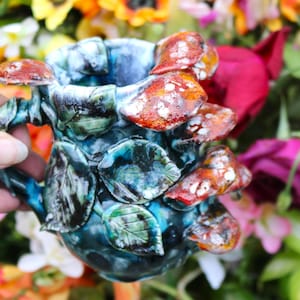 May include: A ceramic mug with a whimsical design featuring red and white spotted mushrooms and green leaves. The mug has a blue and teal glaze, with a handle and decorative elements. The background is blurred with colorful flowers.