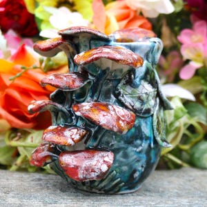 May include: A ceramic vase with a mushroom design. The vase is a deep teal color with red and white mushrooms. The mushrooms are clustered around the vase, with a few green leaves. The vase is on a gray surface.