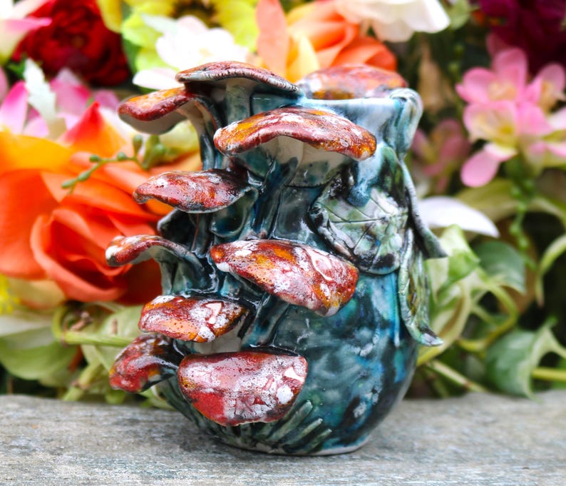 May include: A ceramic vase with a mushroom design. The vase is a deep teal color with red and white mushrooms. The mushrooms are clustered around the vase, with a few green leaves. The vase is on a gray surface.