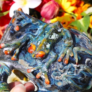May include: A blue and green ceramic mug featuring a frog design. The frog is painted in shades of yellow, orange, and red. The mug has a handle and is decorated with white speckles.