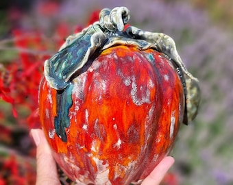 Può includere: Una scultura di zucca in ceramica con una smaltatura lucida e multicolore nei toni dell'arancione vibrante, rosso intenso e bianco. La parte superiore presenta un gambo scolpito e ricurvo e foglie verdi testurizzate con accenti blu scuro.
