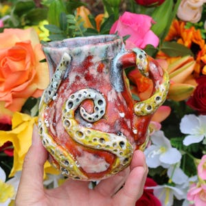 Pode incluir: Uma caneca de cerâmica artesanal com um design único de tentáculo de polvo. A caneca apresenta um esquema de cores vermelho, laranja e amarelo com uma superfície texturizada. A alça e os tentáculos são detalhados com pequenos padrões circulares.